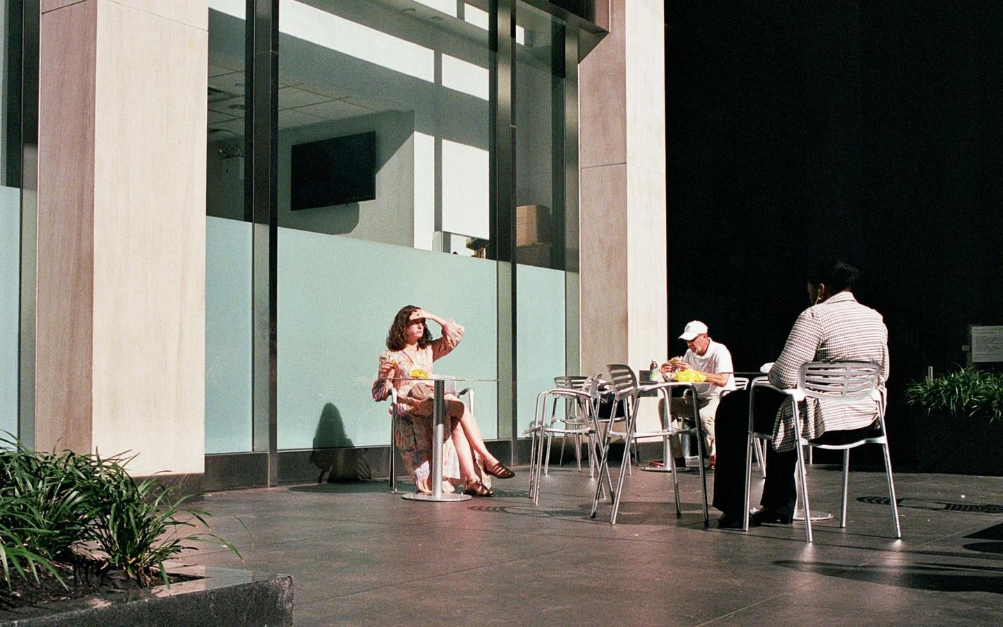 A woman shields her eyes from the sun while sitting at an outdoor table with two men.