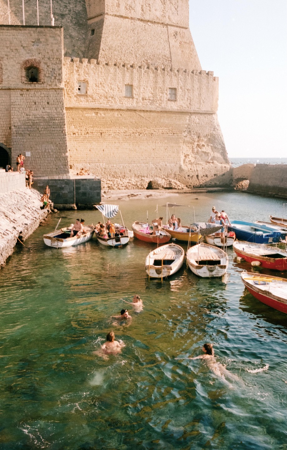 People swim and relax in small boats in a harbor next to a large stone fortress.