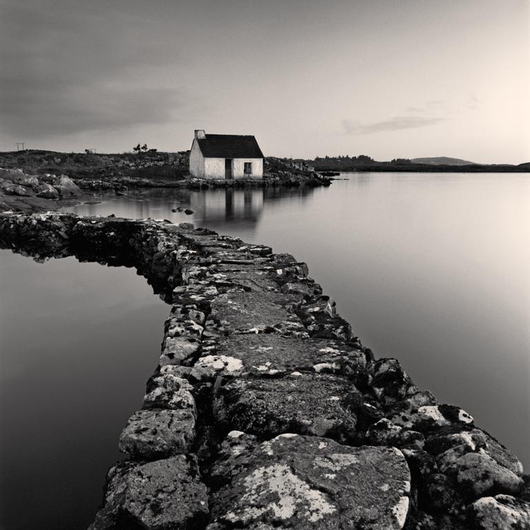 A stone path leads to a small house on the shore of a calm lake under a cloudy sky.