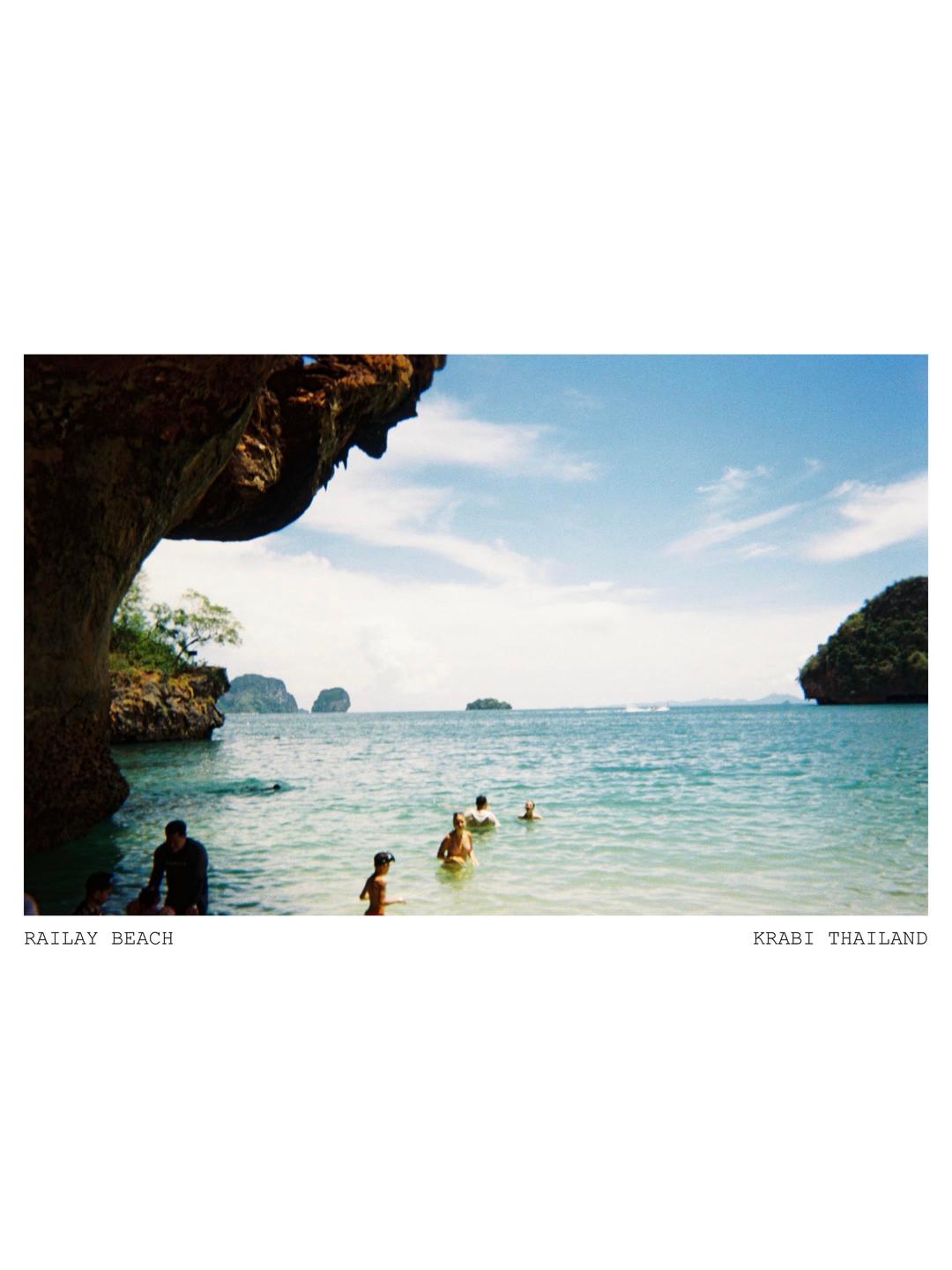 A group of people are swimming in the ocean near a rocky cliff and distant islands under a blue sky.