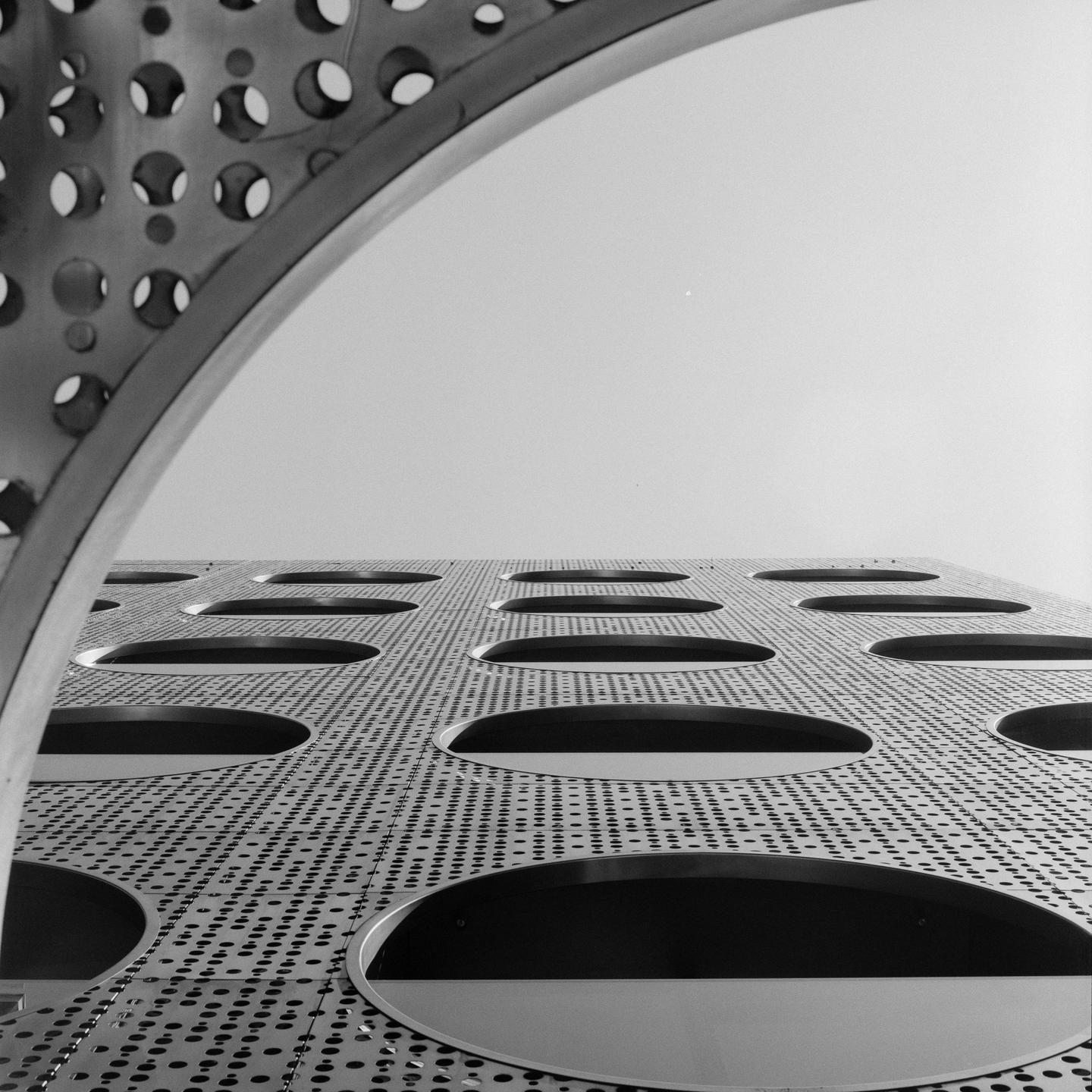 A low-angle view shows a building facade with many small holes and large oval openings, with a curved perforated structure in the foreground.