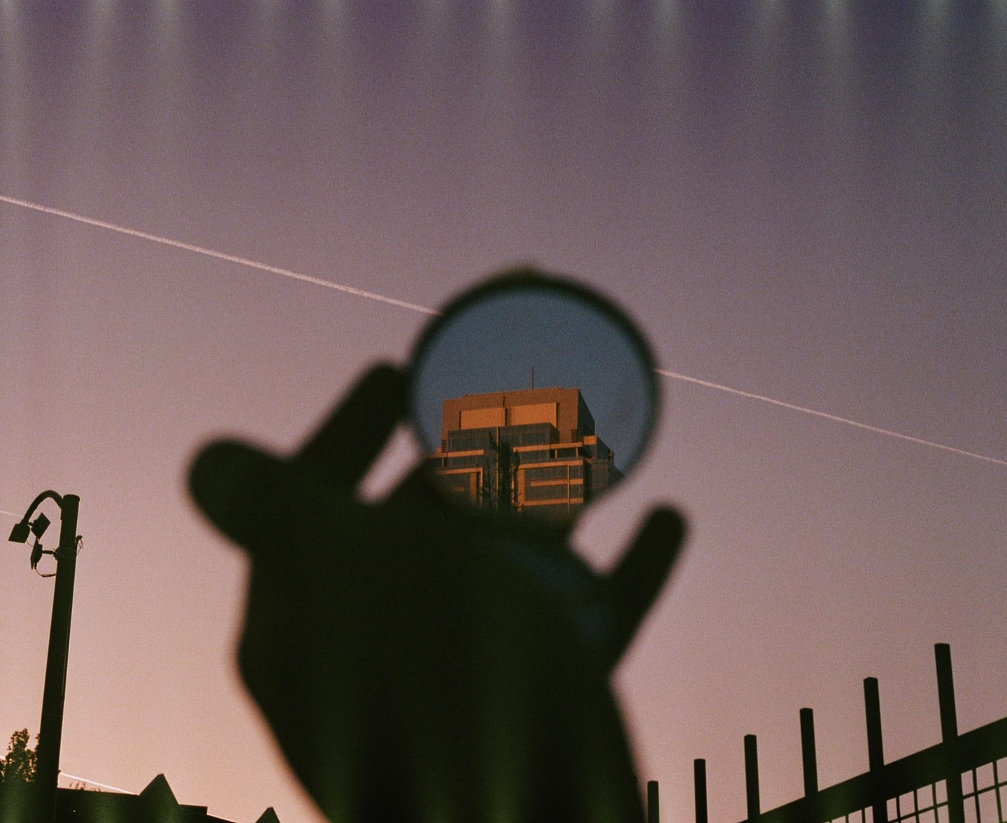 A hand holds a circular mirror reflecting a building against a purple sky with a contrail.