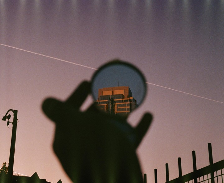 A hand holds a circular mirror reflecting a building against a purple sky with a contrail.