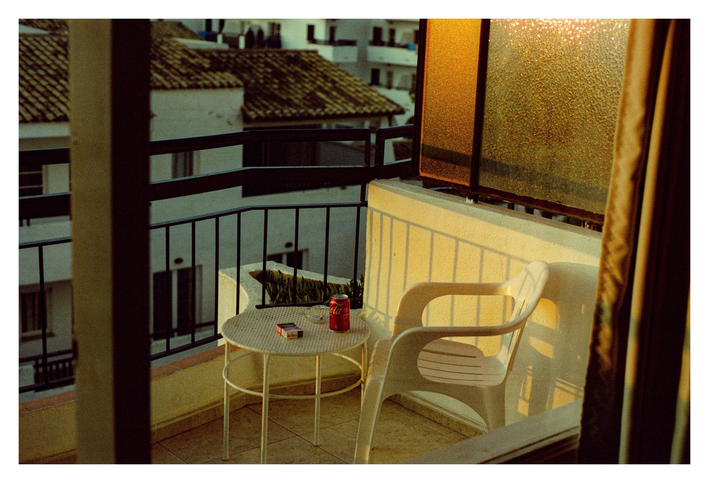 A balcony with a table, chair, and a can of soda and cigarettes on the table, overlooks a town with tiled roofs.