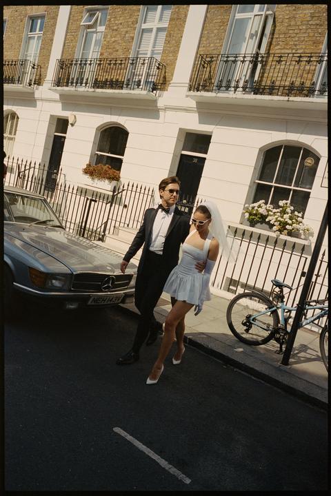 A couple in wedding attire walks along a city street next to a parked car.