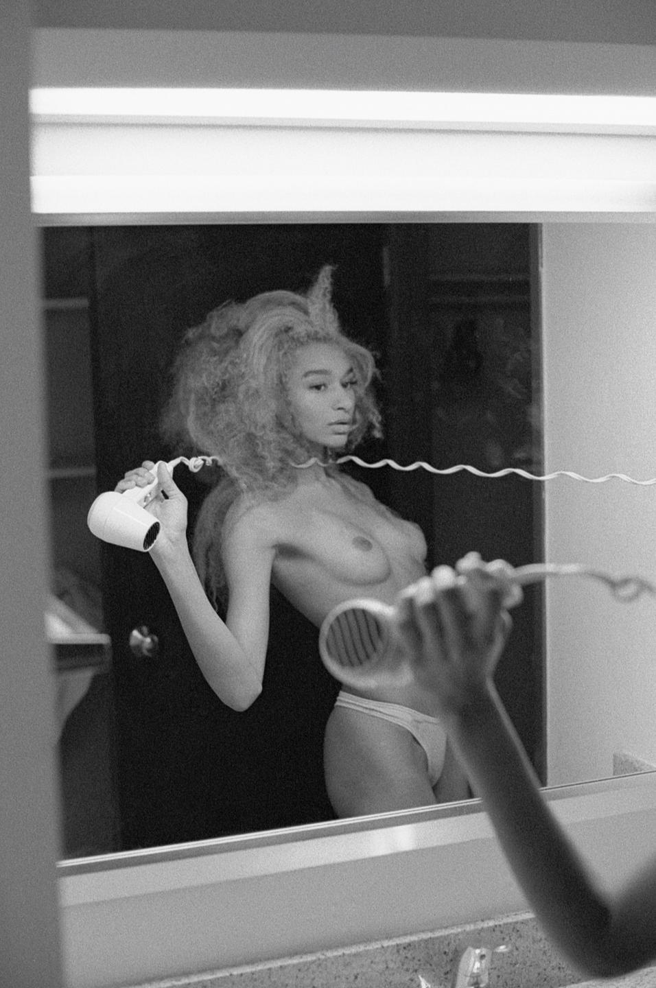 A woman with curly hair holds a hairdryer in front of a mirror, with her reflection visible.