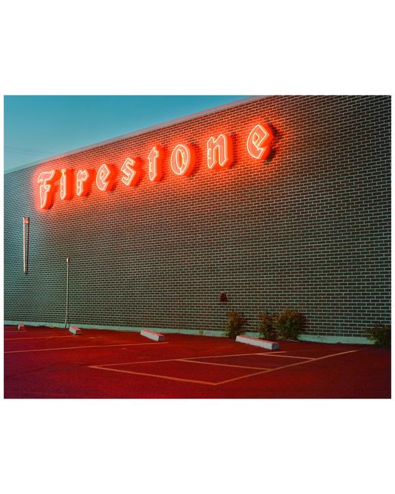 A neon "Firestone" sign glows red above a parking lot at dusk.