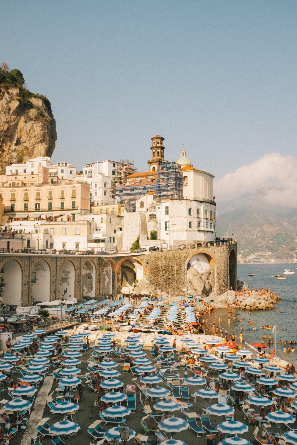 A crowded beach with blue and white striped umbrellas sits below a town built into a cliffside next to the ocean.