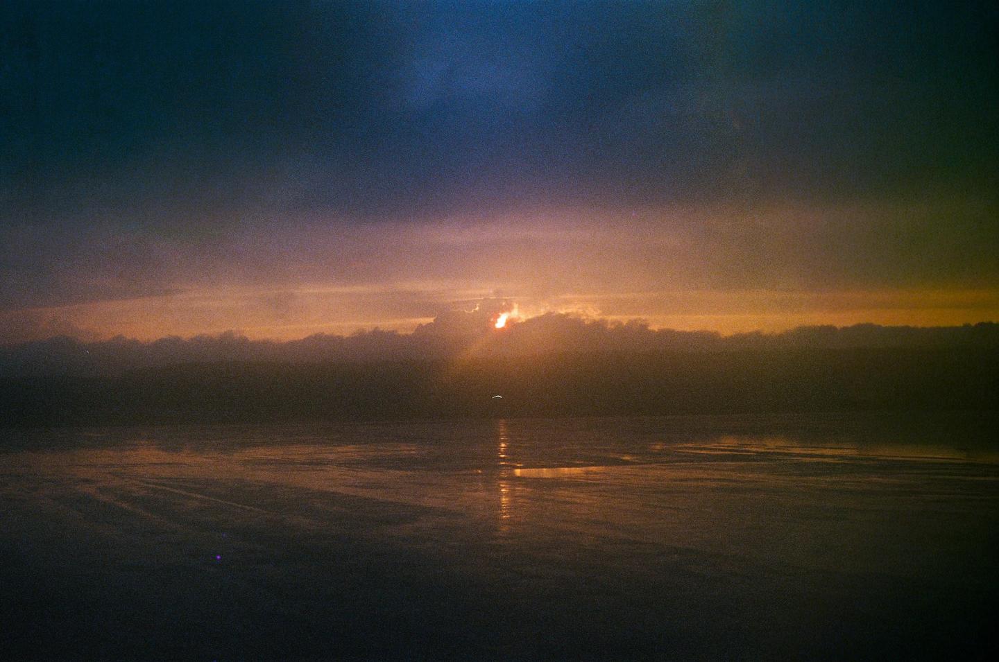 The sun sets behind a cloud bank over a body of water, casting reflections on the surface.