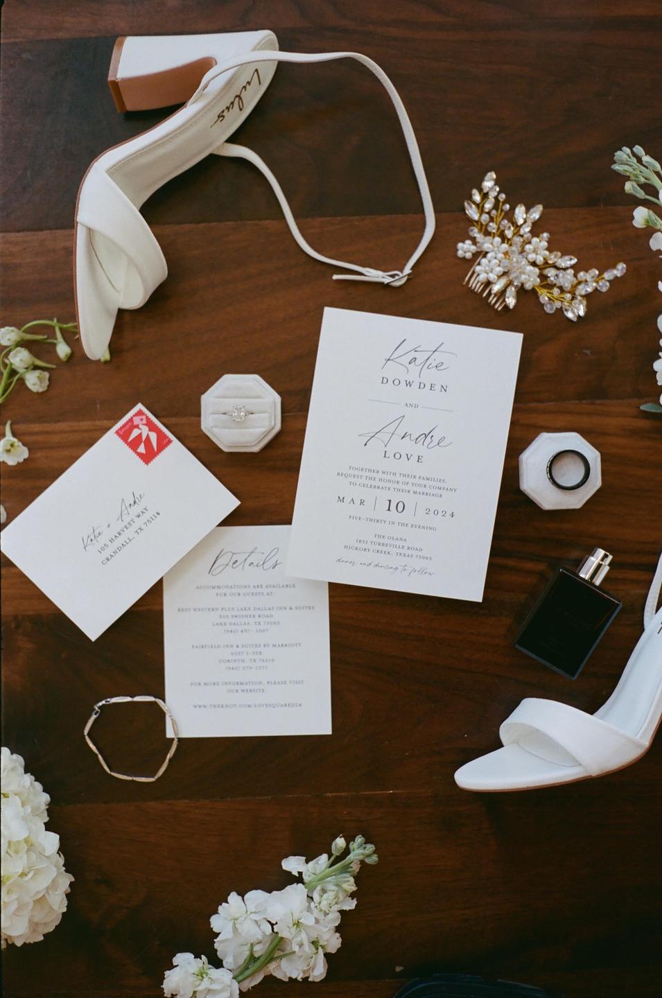 A flat lay shows wedding invitations, a white high heel, a ring, a bracelet, a hair clip, and a perfume bottle on a wooden surface.