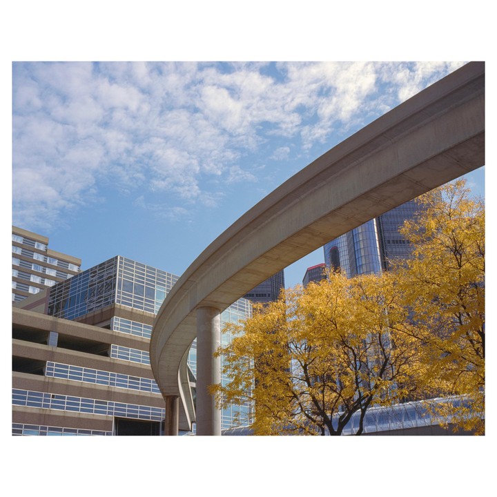 A monorail track curves through a city with buildings and trees with yellow autumn foliage under a blue sky with clouds.