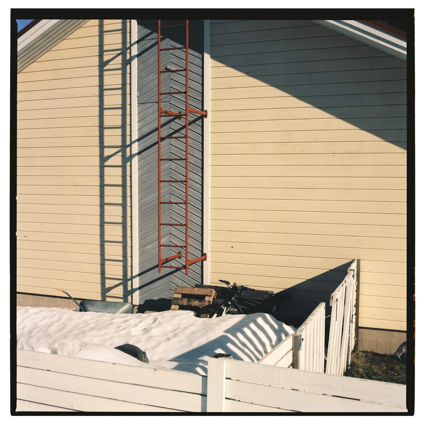 A red ladder is mounted on the side of a yellow house with snow on the ground and a white fence in the foreground.