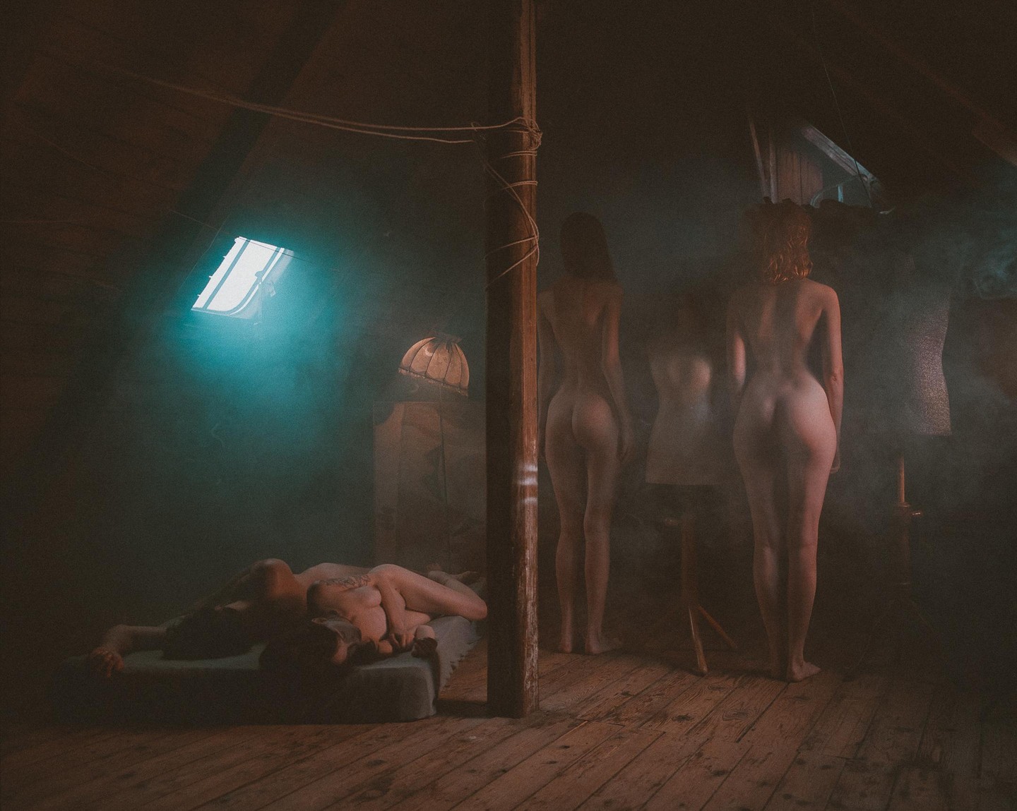Two women stand in a hazy attic with a couple embracing on a mattress and a skylight illuminating the scene.
