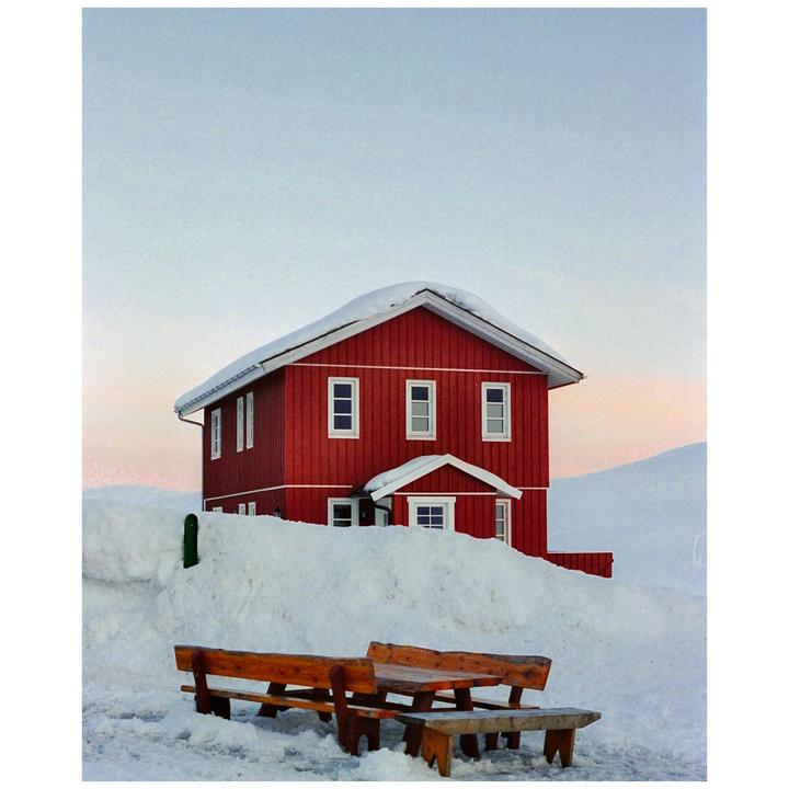 A red house with a snow-covered roof stands in a snowy landscape with wooden benches in the foreground.
