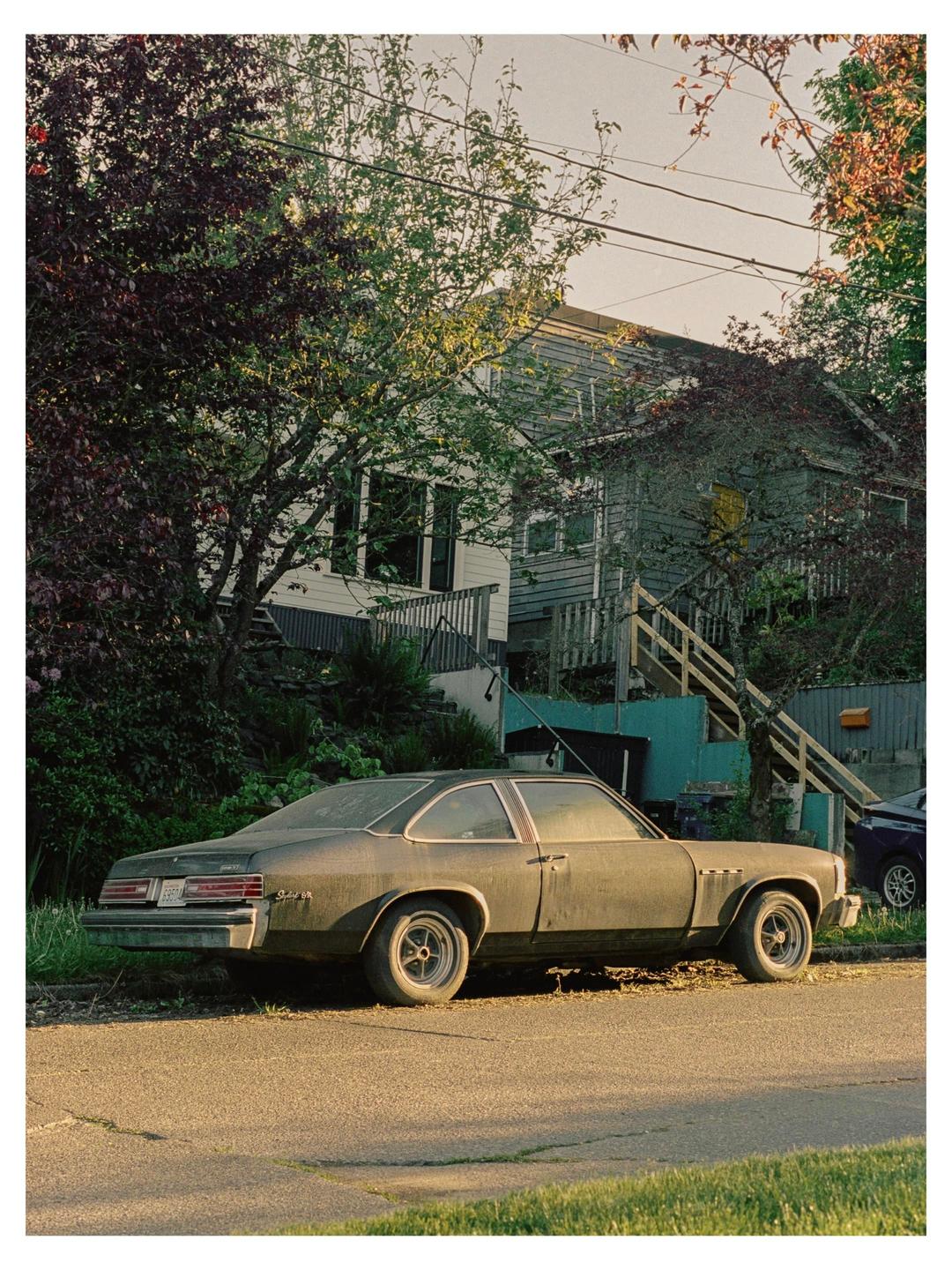 A dusty car is parked on the side of a street in front of a house with trees and power lines.