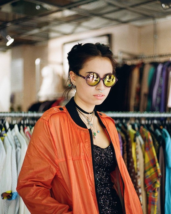 A woman wearing an orange jacket and round sunglasses stands in front of a rack of clothes.