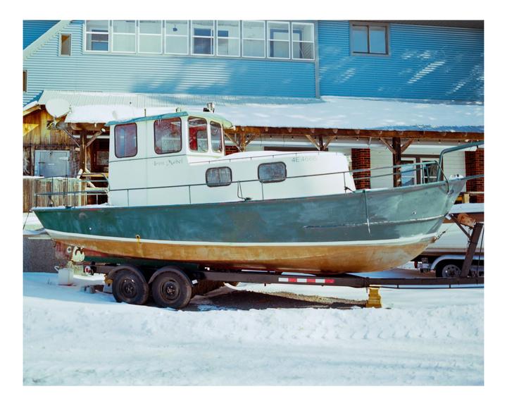 A boat named "Iron Mabel" sits on a trailer in the snow in front of a building.
