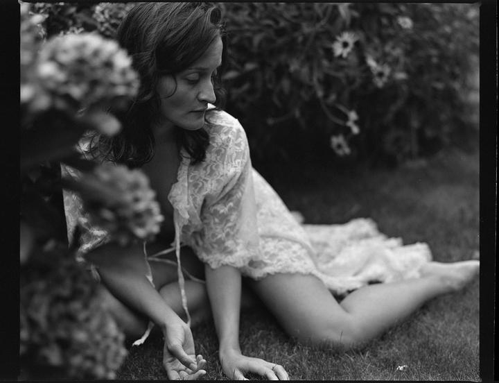 A woman in a lace garment is sitting on the grass with flowers in the foreground and background.
