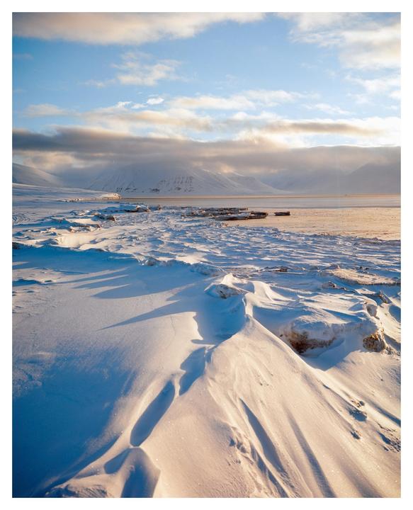 Snow drifts cover the ground with mountains and a body of water in the background under a cloudy sky.