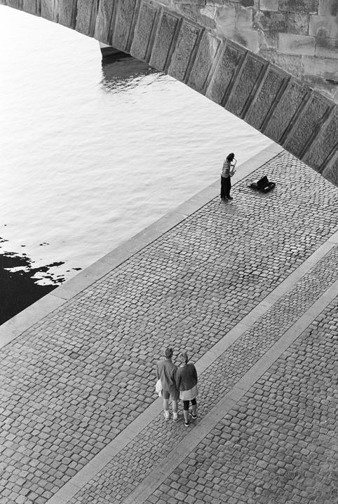 A musician plays a saxophone on a cobblestone riverbank under a bridge, while a couple walks nearby.