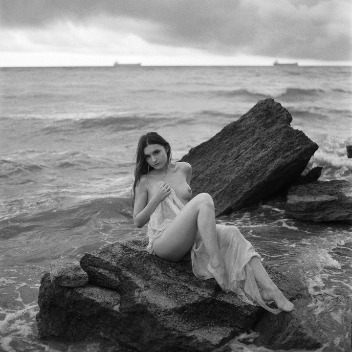 A woman sits on a rock in the sea, partially covered by sheer fabric, with ships visible on the horizon under a cloudy sky.