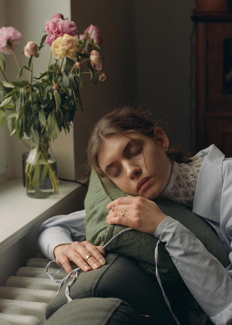 A woman sleeps with her head on a green pillow next to a window with a vase of flowers.