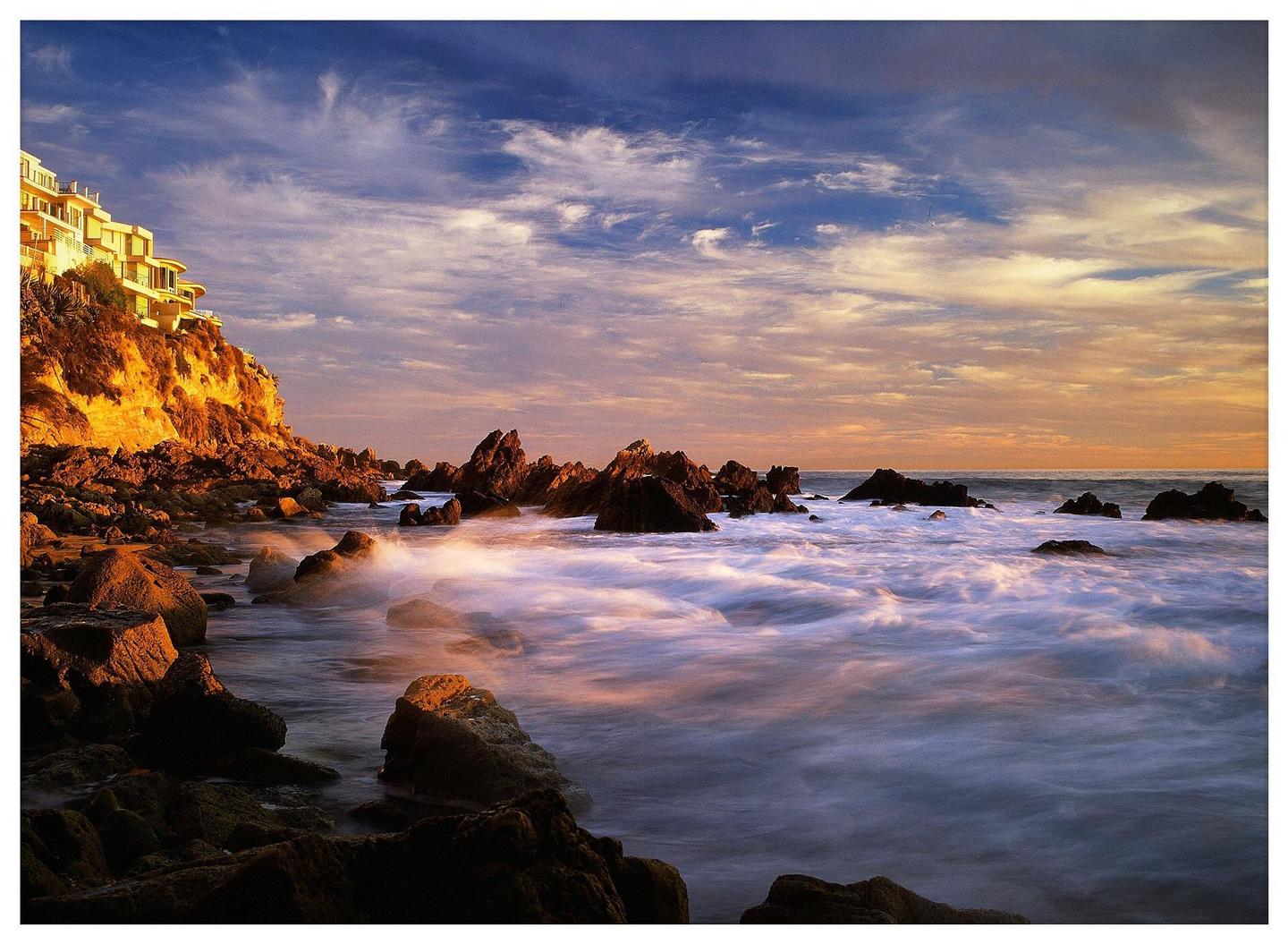 A building sits atop a cliff overlooking a rocky coastline with waves crashing against the shore under a dramatic sky.