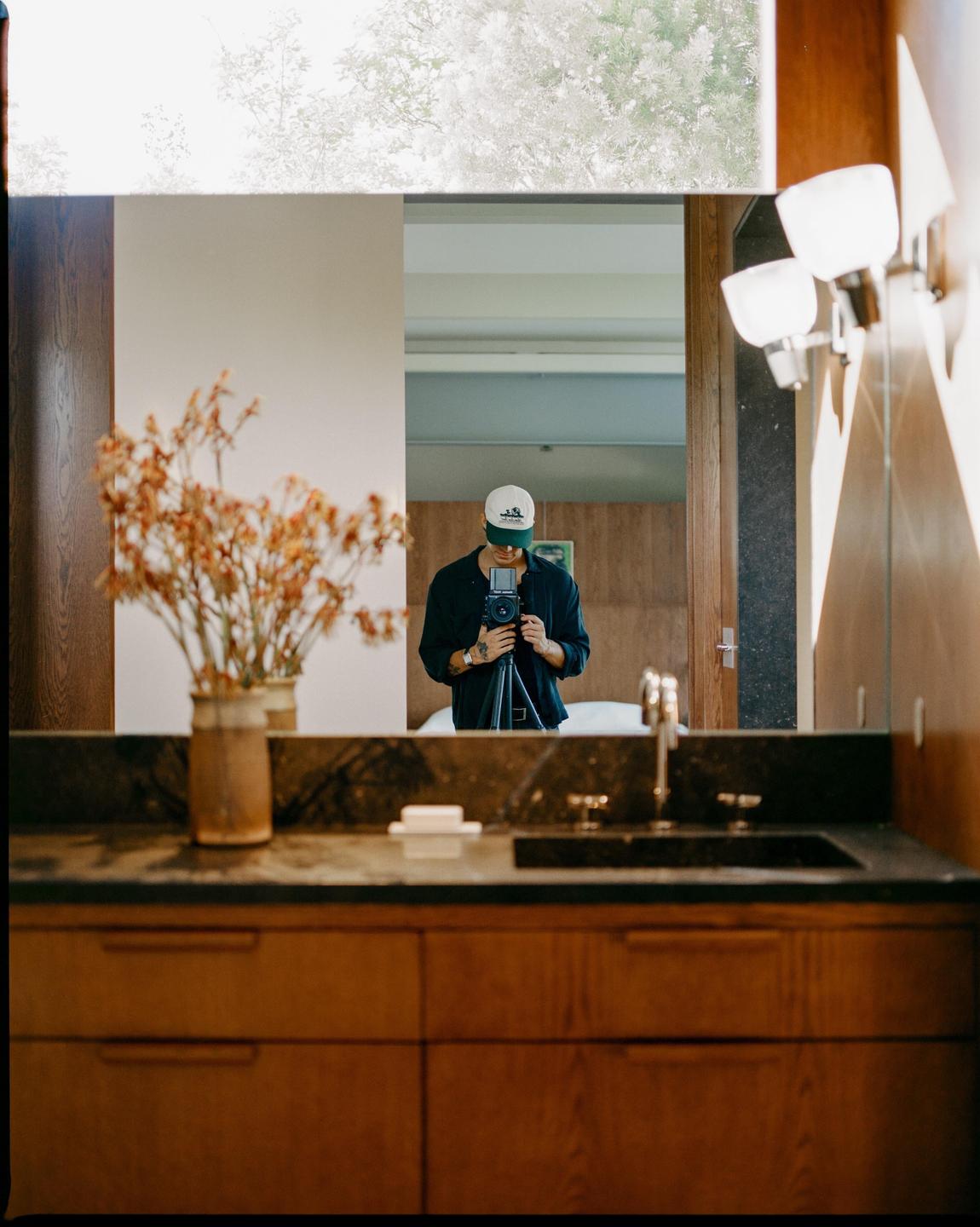A man wearing a cap and holding a camera on a tripod is reflected in a bathroom mirror.