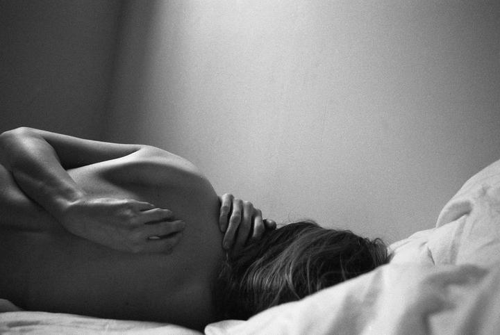A woman lies on her side on a bed with her head turned away from the viewer, her hands clasped behind her neck.