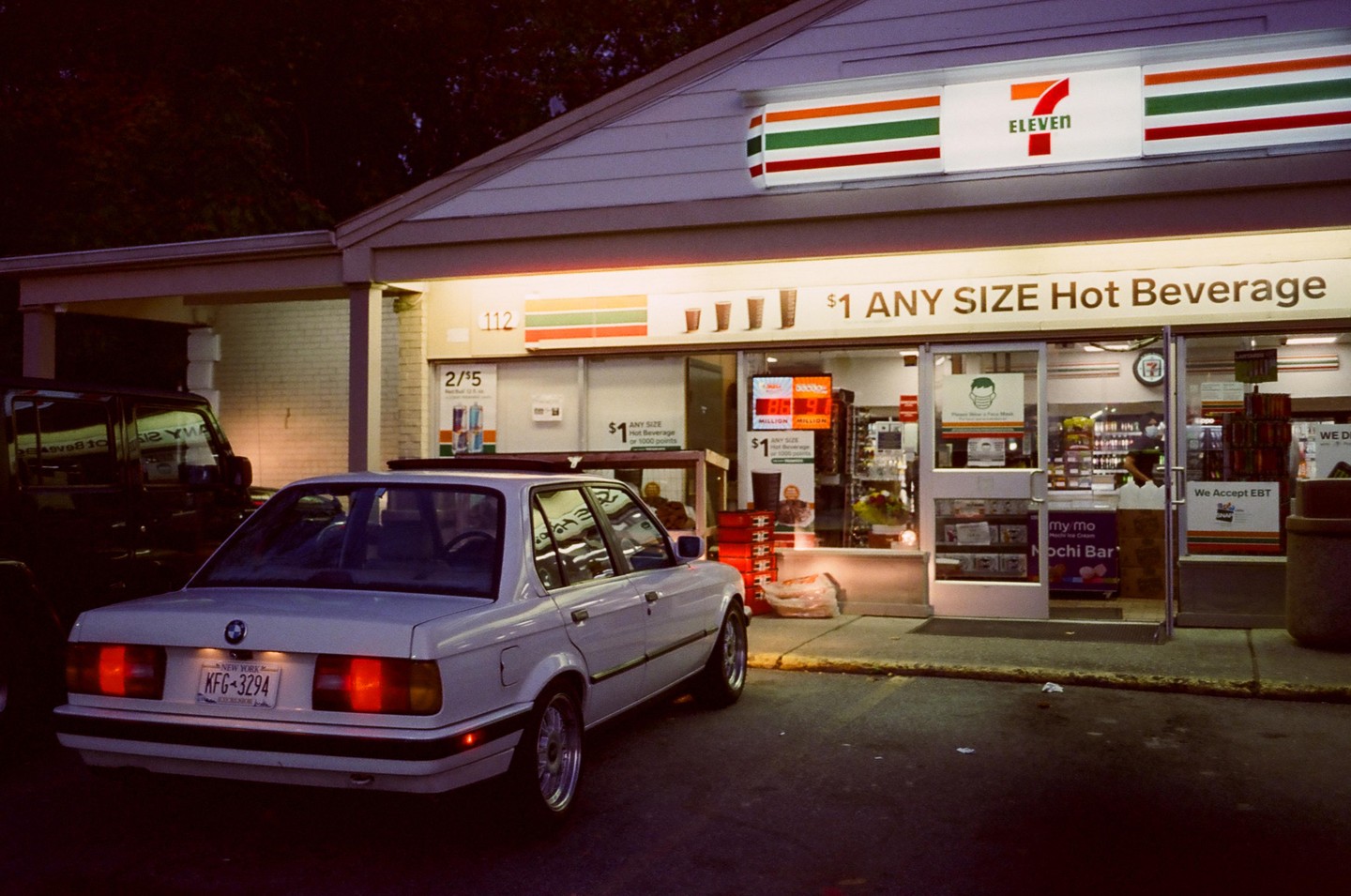 A white sedan is parked in front of a brightly lit 7-Eleven convenience store at dusk.