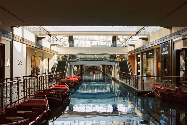 A canal with red boats runs through a modern shopping mall with multiple levels and glass railings.