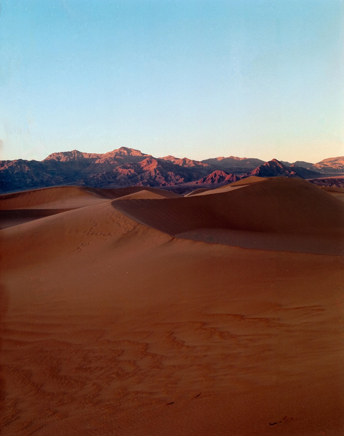 Sand dunes stretch across the foreground with mountains in the distance under a clear sky.