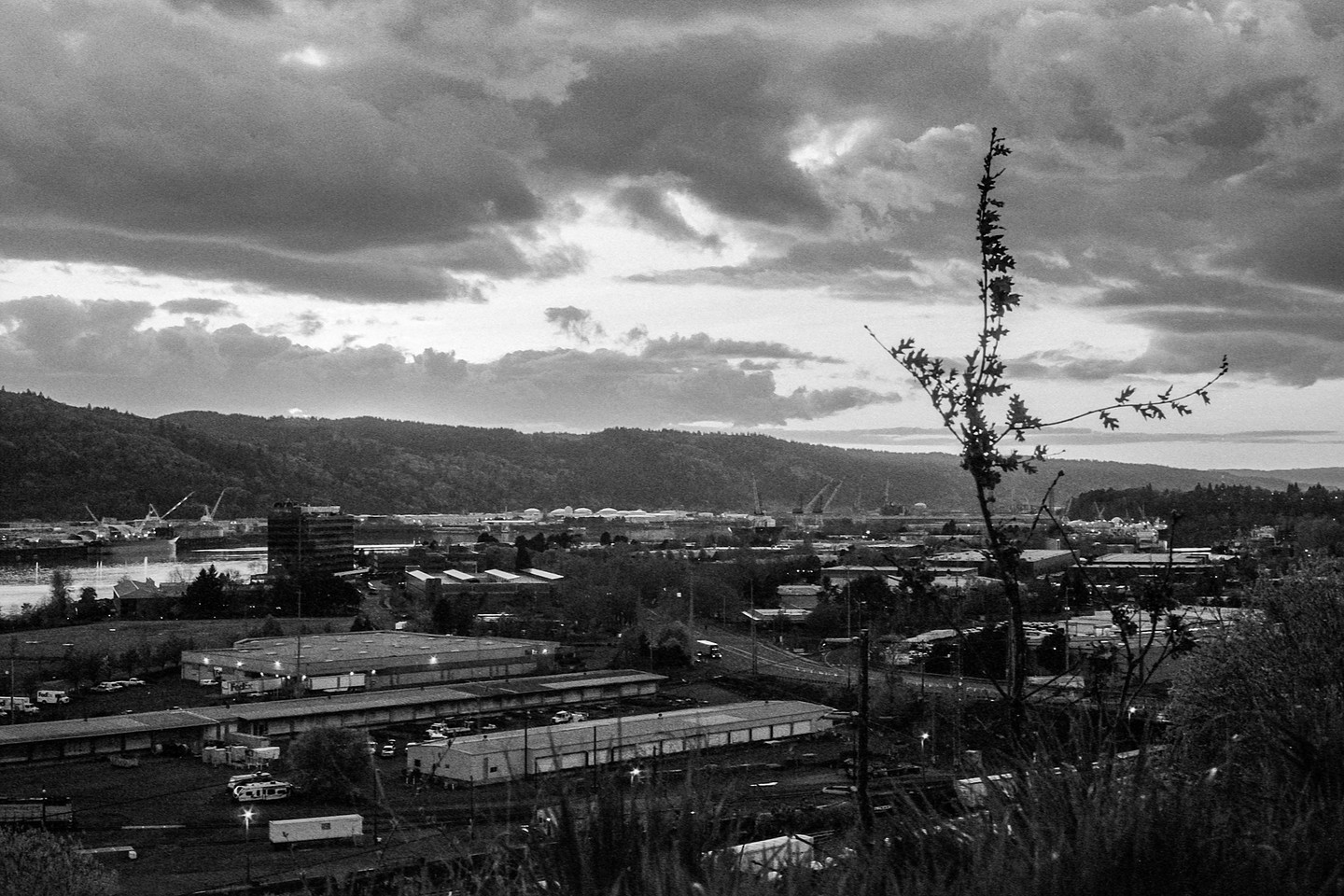 A black and white landscape shows a city with industrial buildings and a river, backed by hills under a cloudy sky.