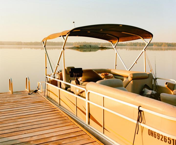 A pontoon boat is docked at a wooden pier on a calm lake with a forested shore in the distance.