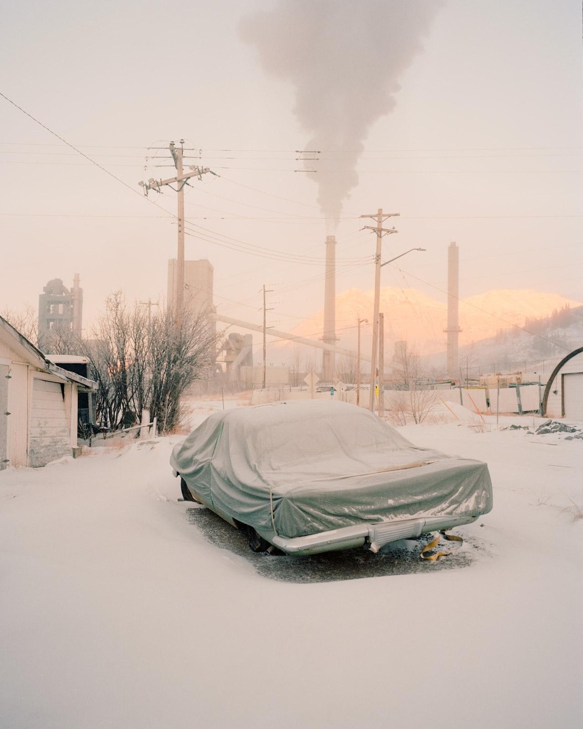 A car covered with a tarp is parked in the snow with industrial buildings and mountains in the background.