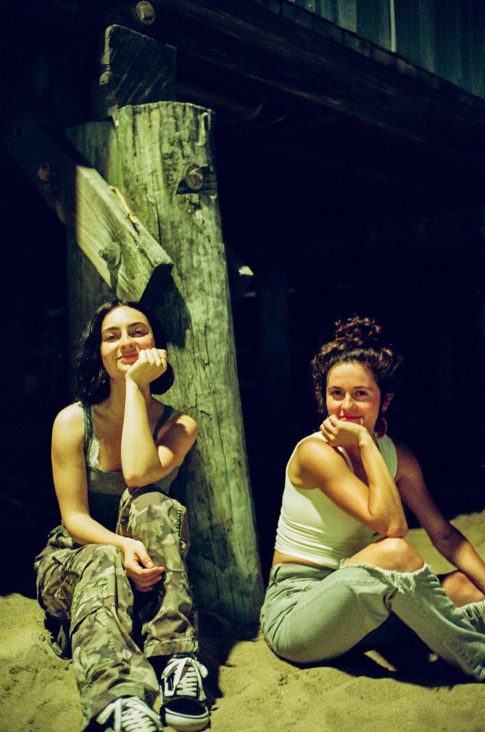 Two women sit on sand under a pier, illuminated by a bright light source.