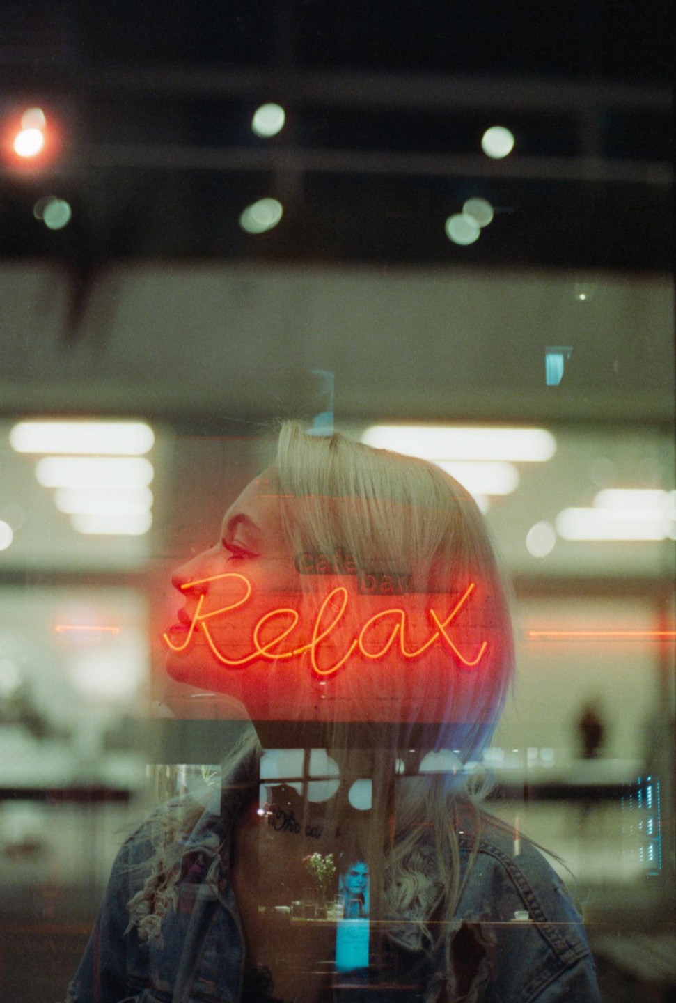 A woman with blonde hair is visible through a window with a neon "Relax" sign reflected on it.