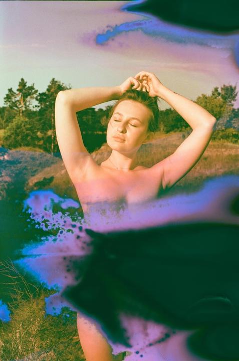 A woman with her eyes closed and hands behind her head stands in a field with trees in the background, with purple and blue film soup effects.