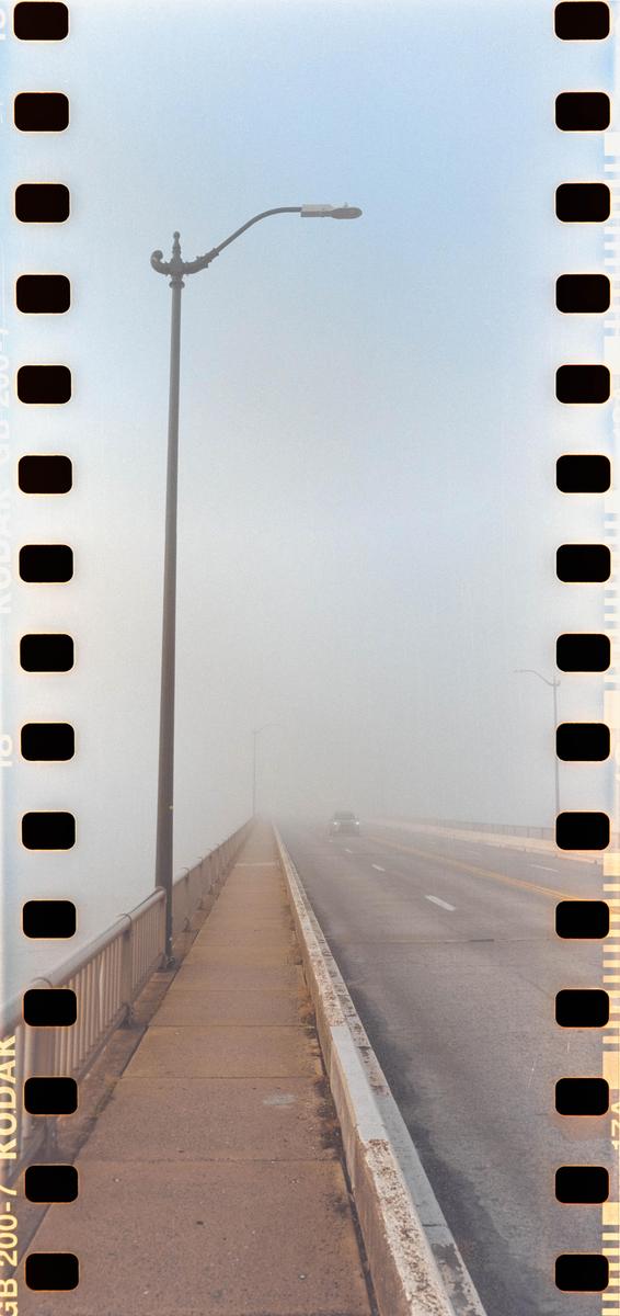 A car drives on a foggy bridge with a street light on the left.
