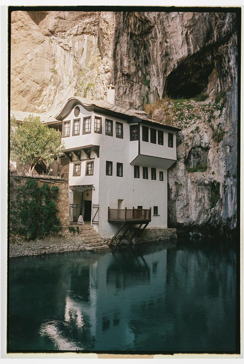 A white house with a balcony and a stone staircase sits beside a body of water, with a large rock face rising behind it.