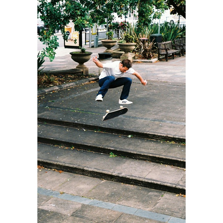 A man performs a kickflip on a skateboard over a set of concrete steps in a park.
