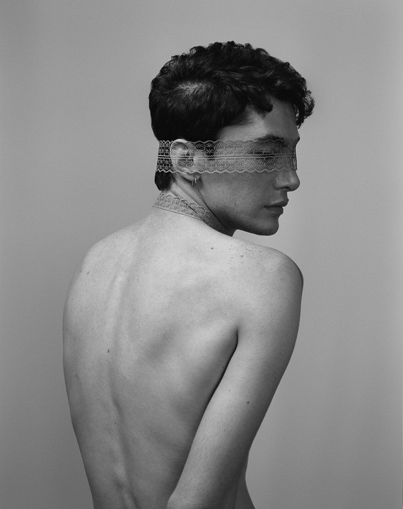 A man with curly hair and a lace blindfold looks over his shoulder, showing his bare back.