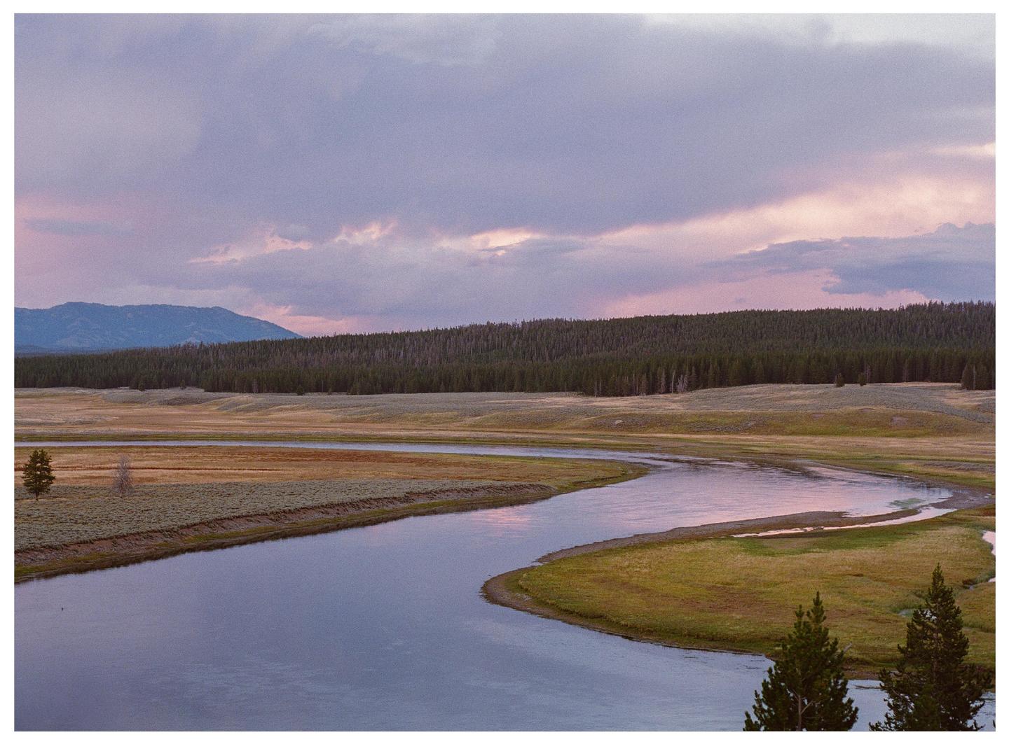 A river winds through a grassy landscape with a forest and mountains under a colorful sky.