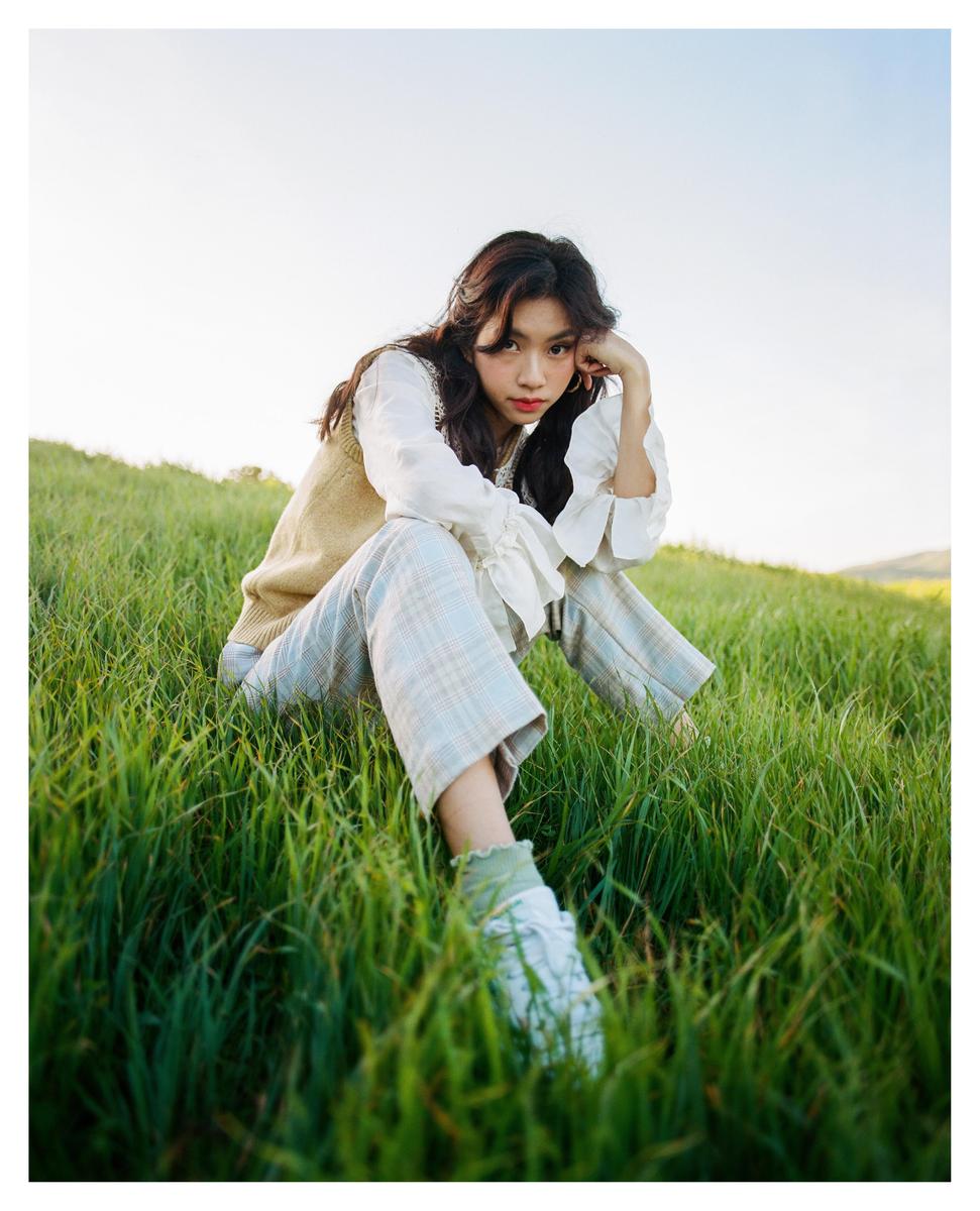 A woman sits in a field of tall green grass with her hand resting on her chin.