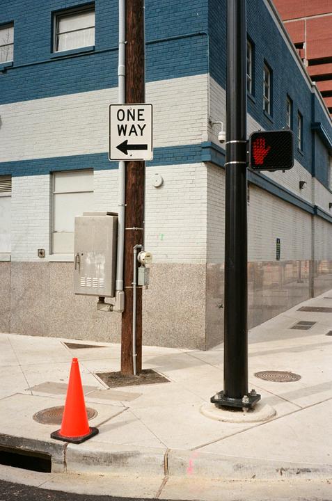 A one way sign is attached to a utility pole on a street corner with a traffic cone on the sidewalk.