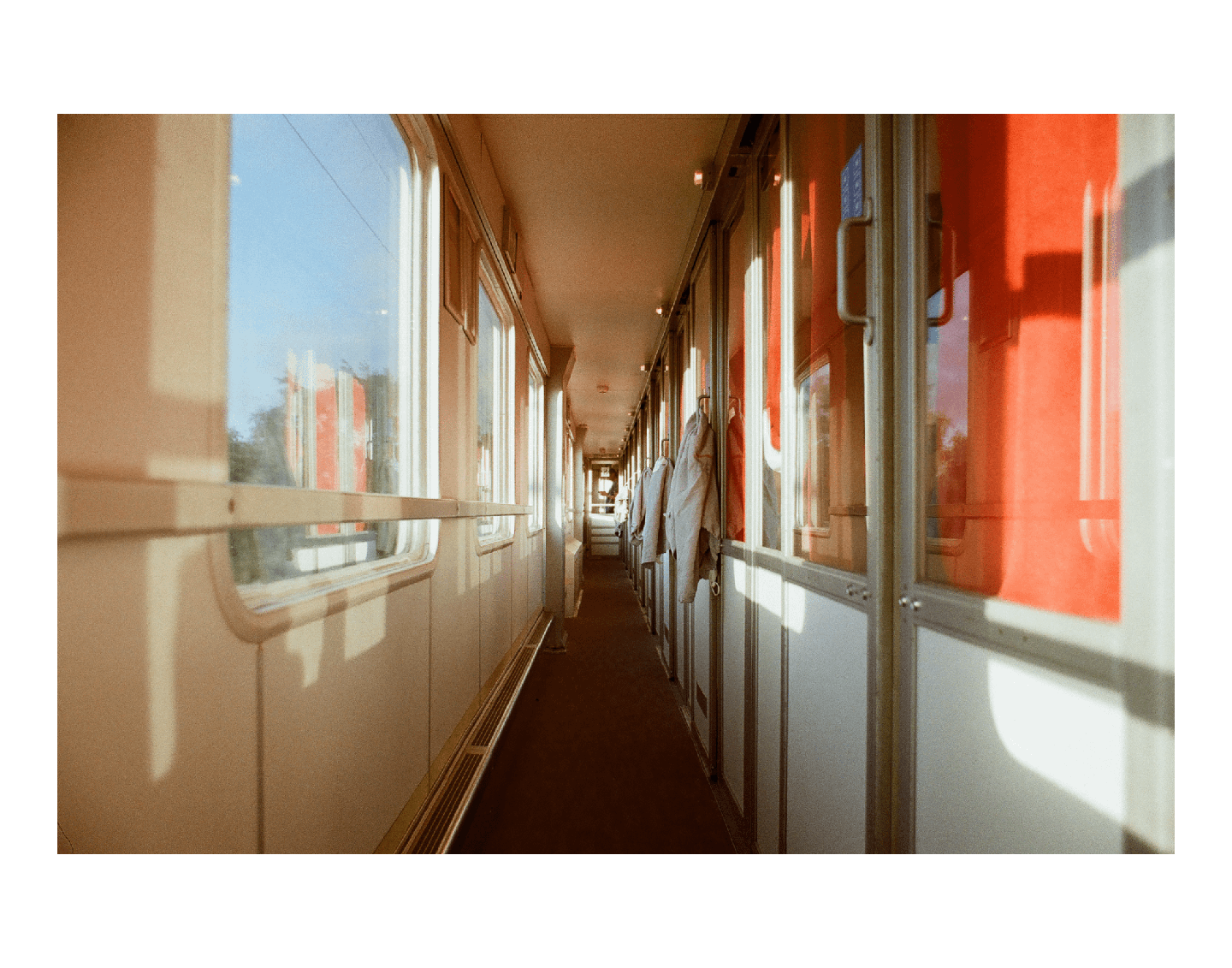 A long, narrow train corridor is visible with windows on the left and compartment doors on the right, some of which reflect a bright red color.