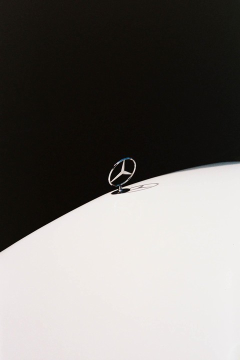 A Mercedes-Benz hood ornament is visible against a stark black background and a white car body.