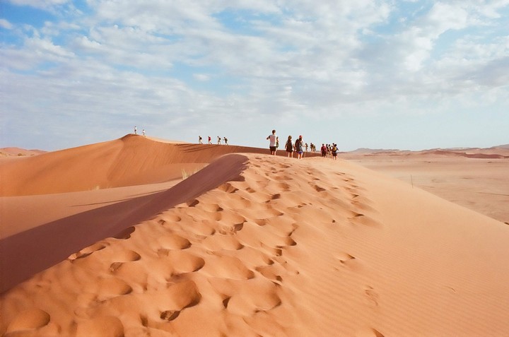 A group of people walk along the ridge of a large sand dune under a partly cloudy sky.