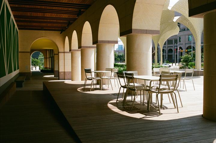 A row of arches and columns extends along a wooden deck with tables and chairs, leading to a city street in the distance.