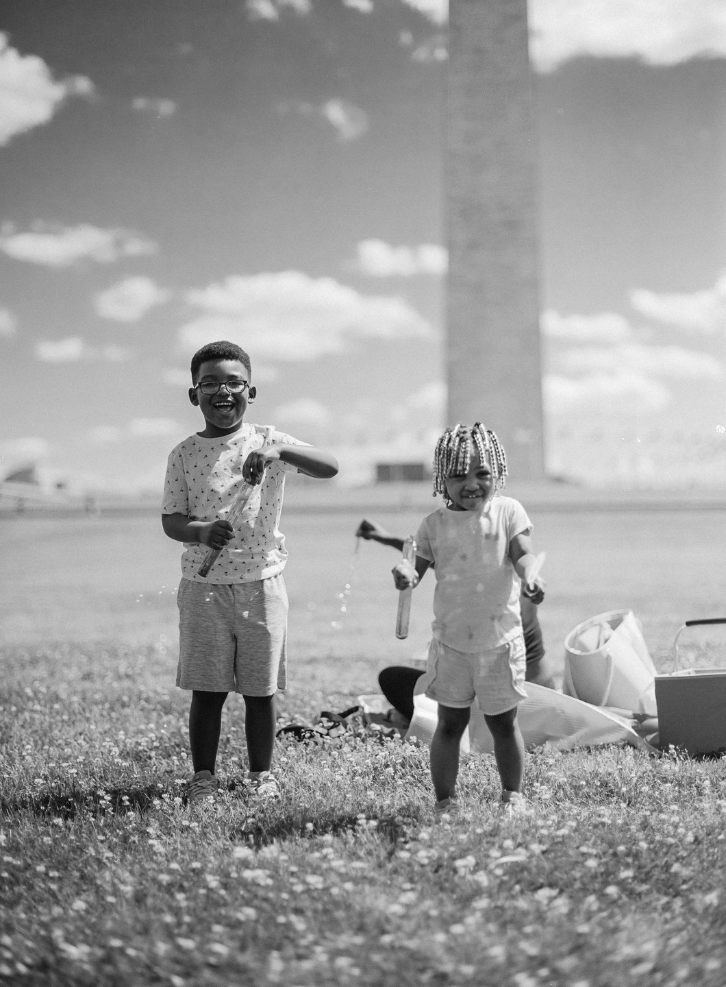 Two children play with bubble wands in a grassy field with a tall monument in the background.
